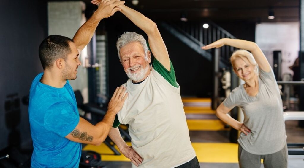 Animateur-aidant-un-senior-homme-à-faire-un-étirement-latéral-lors-d-une-séance-de-gym-douce-en-groupe