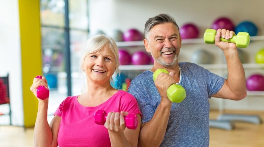Couple-de-seniors-souriant-tenant-des-haltères-lors-d'une-séance-de-gym-douce-en-salle-de-sport