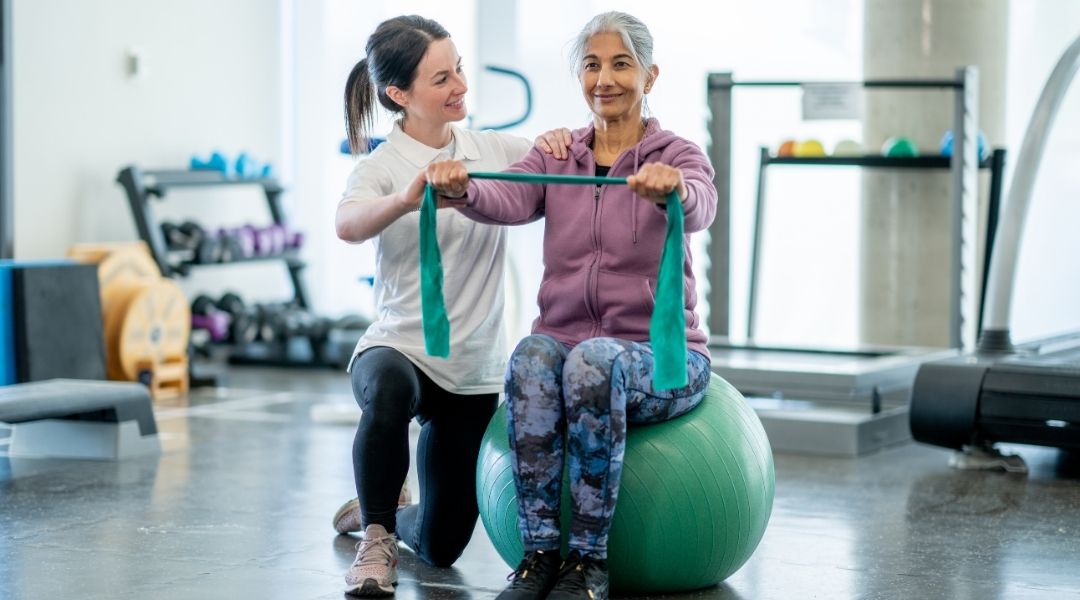 Coach-guidant-une-femme-senior-dans-un-exercice-de-renforcement-avec-élastique-sur-ballon-de-gym-douce